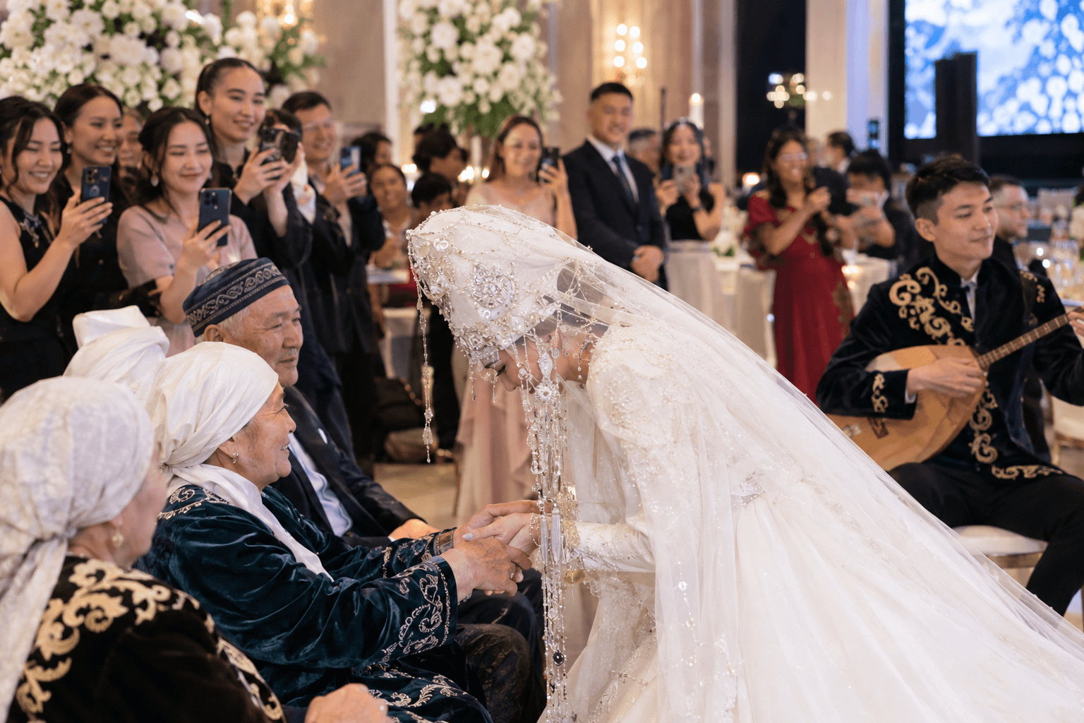 A Kazakh bride in a saukele bows to greet the elders at her wedding while a young man plays the dombra
